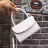 Luxury white leather handbag with textured design and hard handle against a brick wall background.